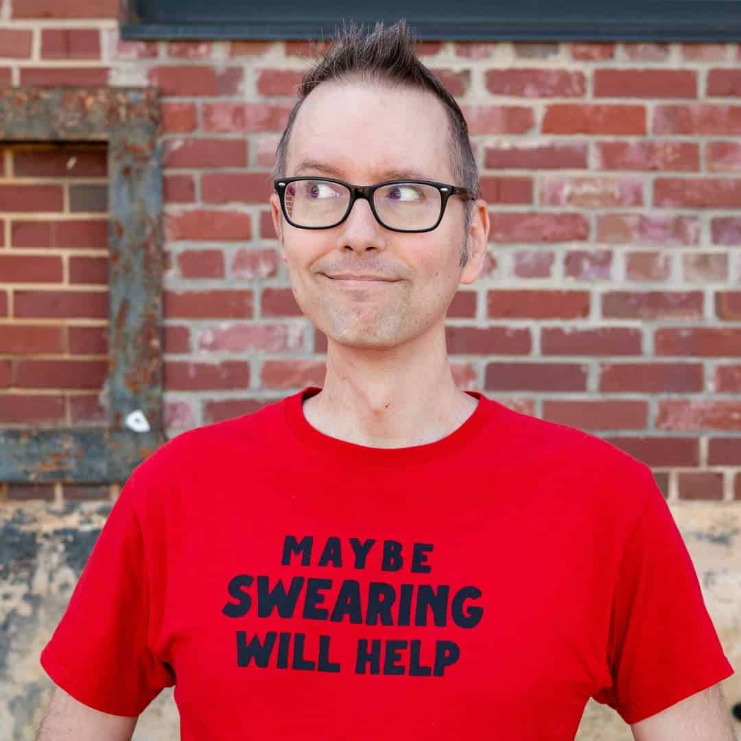 A person with glasses and a red shirt that says "Maybe Swearing Will Help" stands in front of a brick wall, looking to the side and smiling slightly.