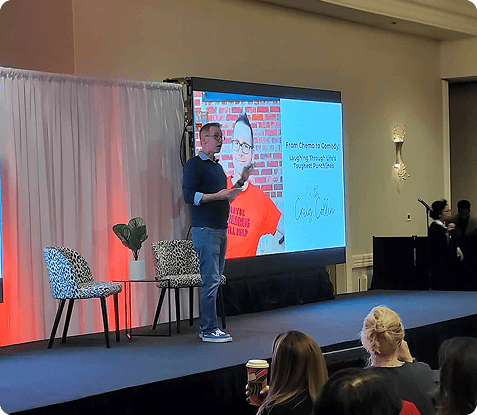 A man stands on stage giving a presentation to an audience, with a slide displayed behind him featuring a photo and text. Two chairs and a small table are on stage.
